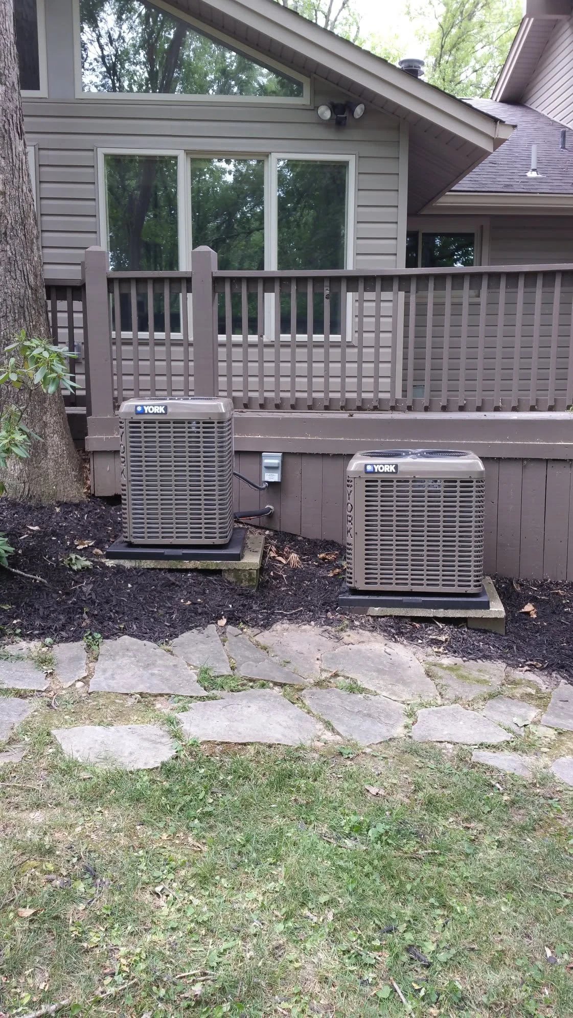 Two York HVAC units installed in front of a brown residential home with a deck and large windows